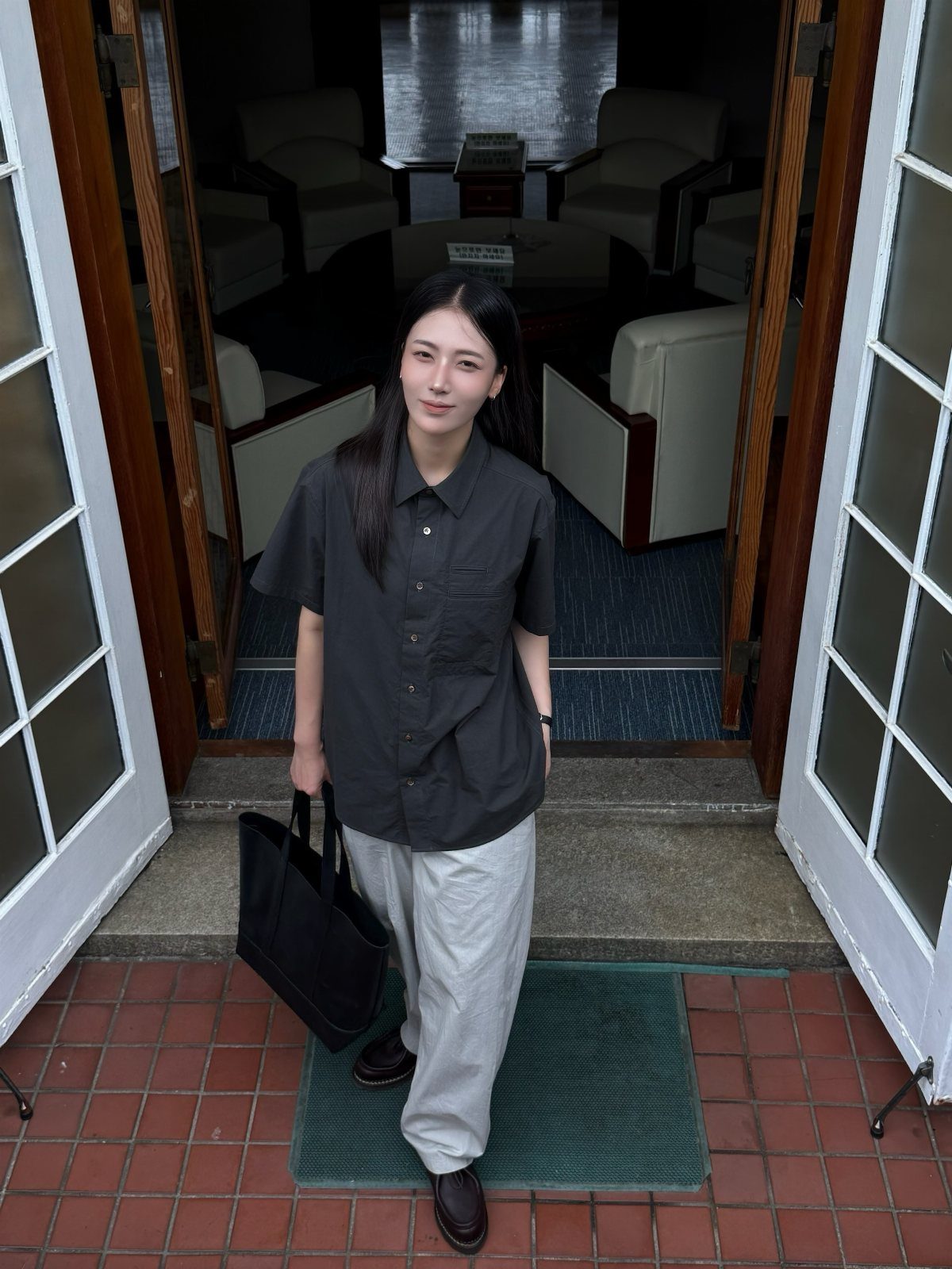 A woman in a dark grey shirt and light grey trousers standing in a doorway holding a black bag.