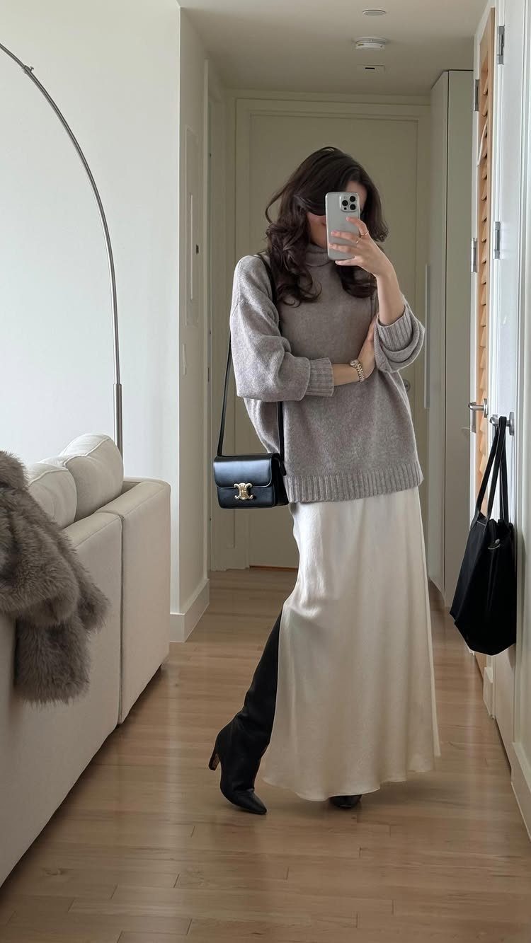 Woman in a grey sweater and cream skirt posing for a mirror selfie in a hallway.