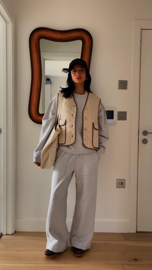 Woman wearing a grey sweatshirt, wide-leg trousers, and a beige utility vest standing indoors.