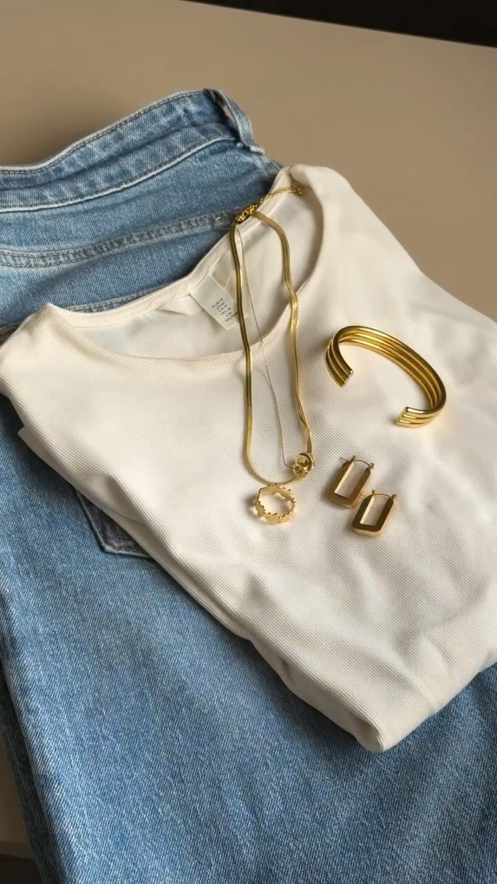White t-shirt and blue jeans paired with gold jewelry on a flat surface.
