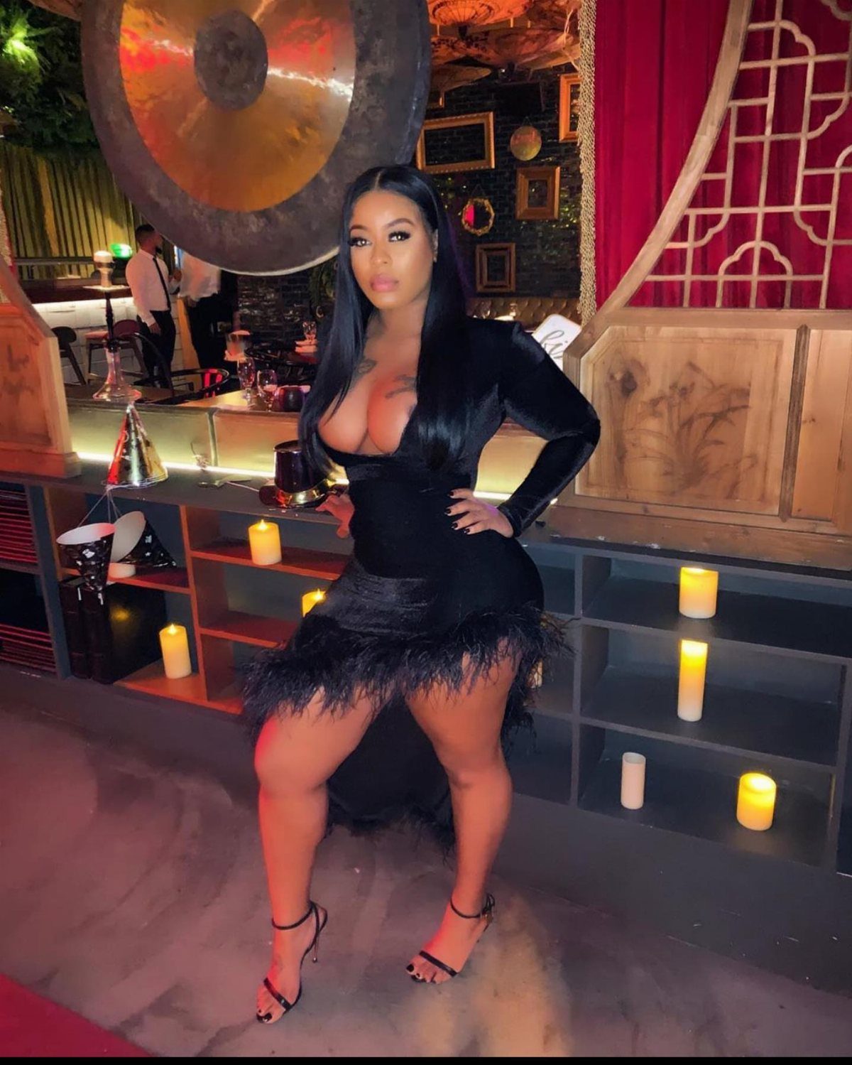 A woman posing in a black feathered mini dress and strappy heels in a lounge.