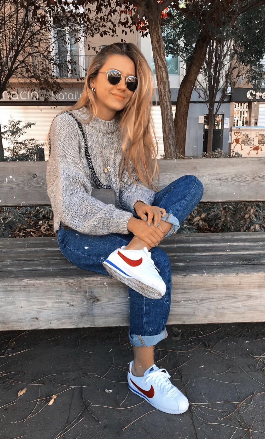 Woman sitting on a wooden bench wearing blue jeans and white Nike Cortez sneakers.
