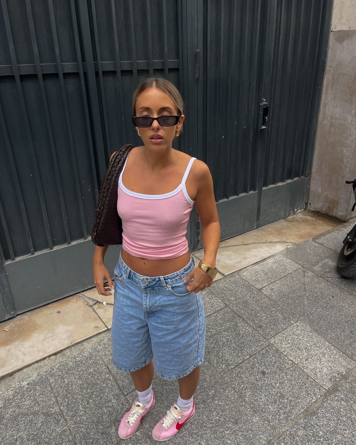 Woman in pink tank top, denim jorts, and pink Nike Cortez sneakers standing by a dark gate.