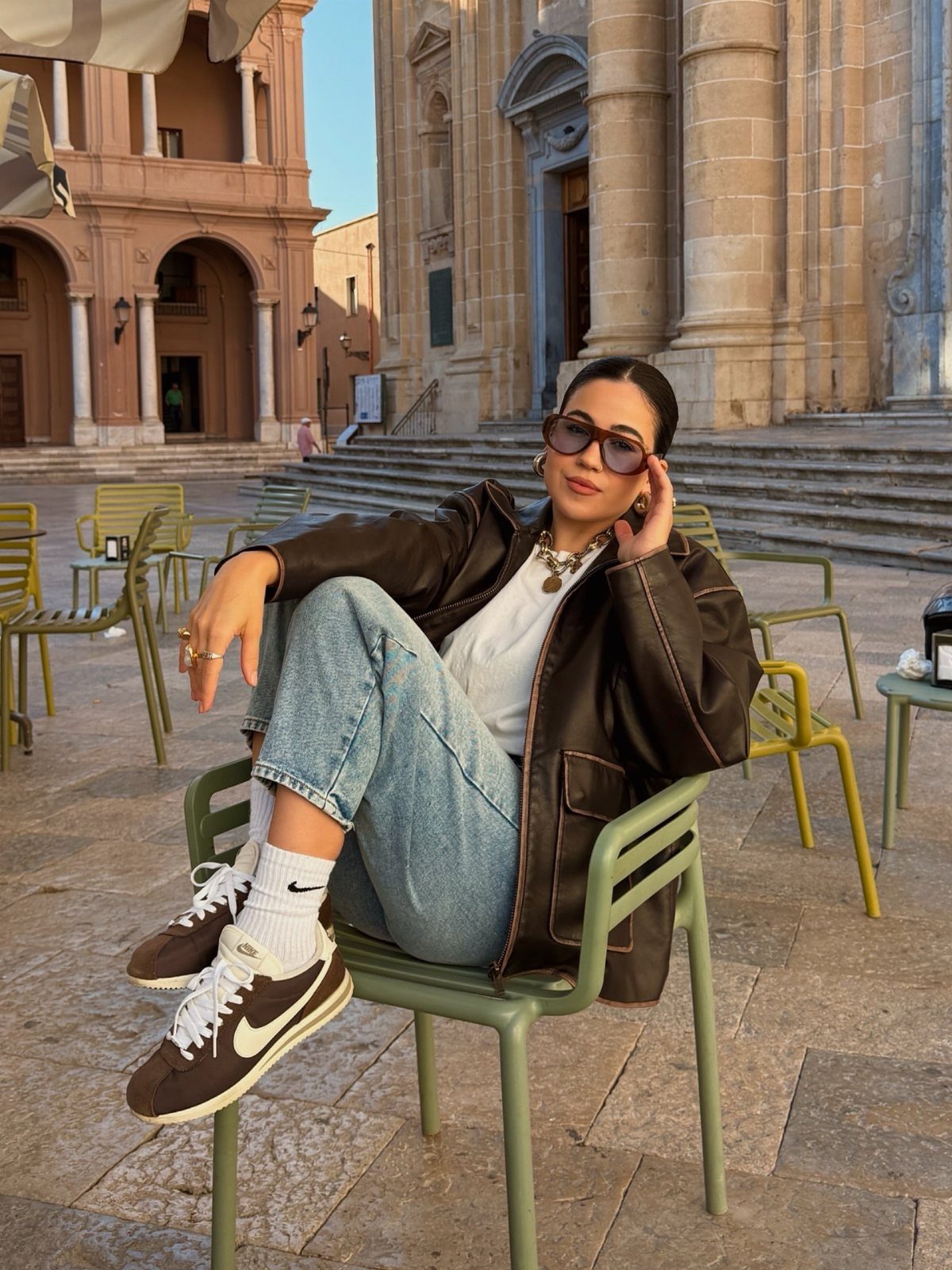 Woman sitting in a chair wearing a brown leather jacket, denim jeans, and brown Nike Cortez sneakers.