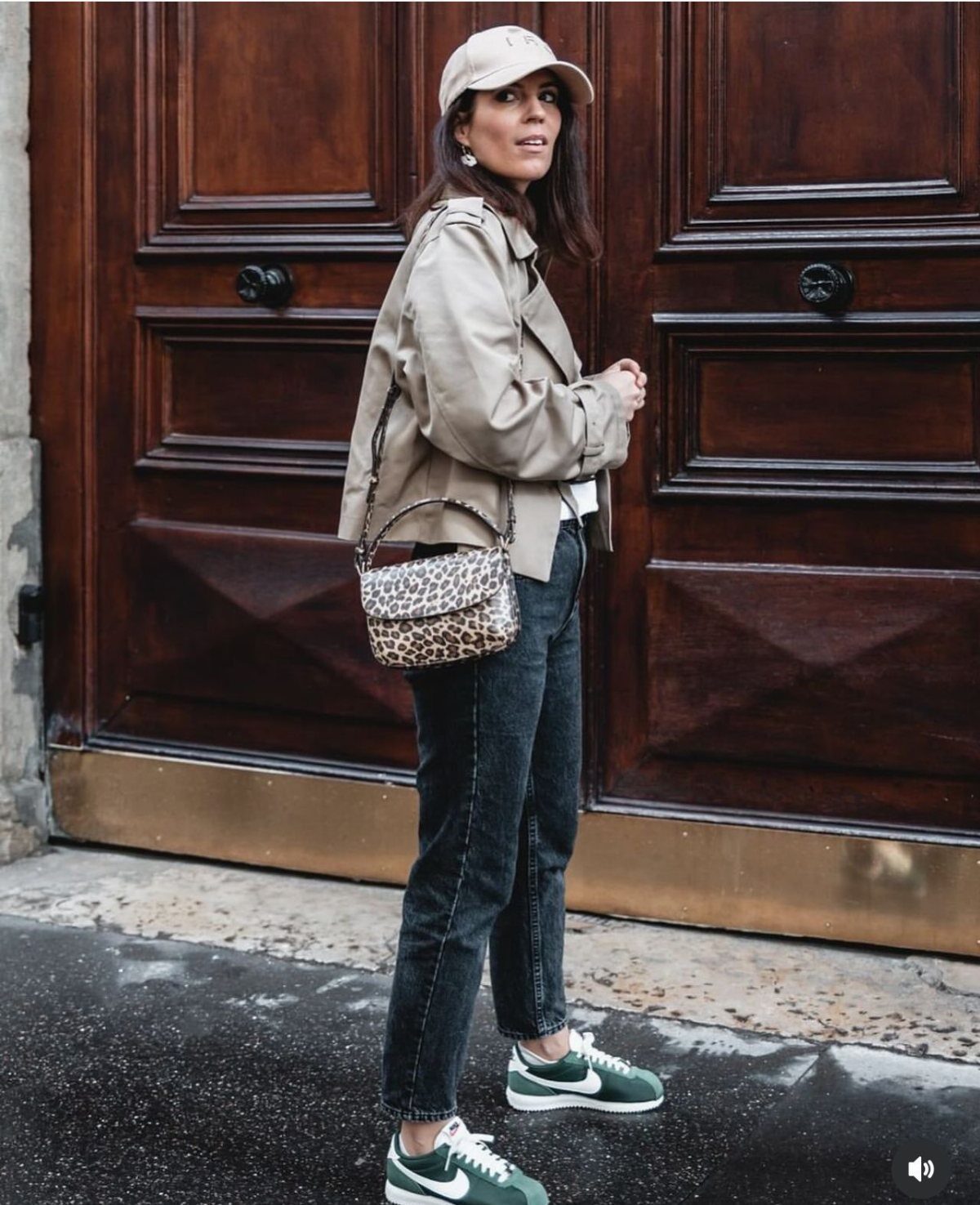 Woman wearing a beige trench coat, dark jeans, and green Nike Cortez sneakers standing by a wooden door.