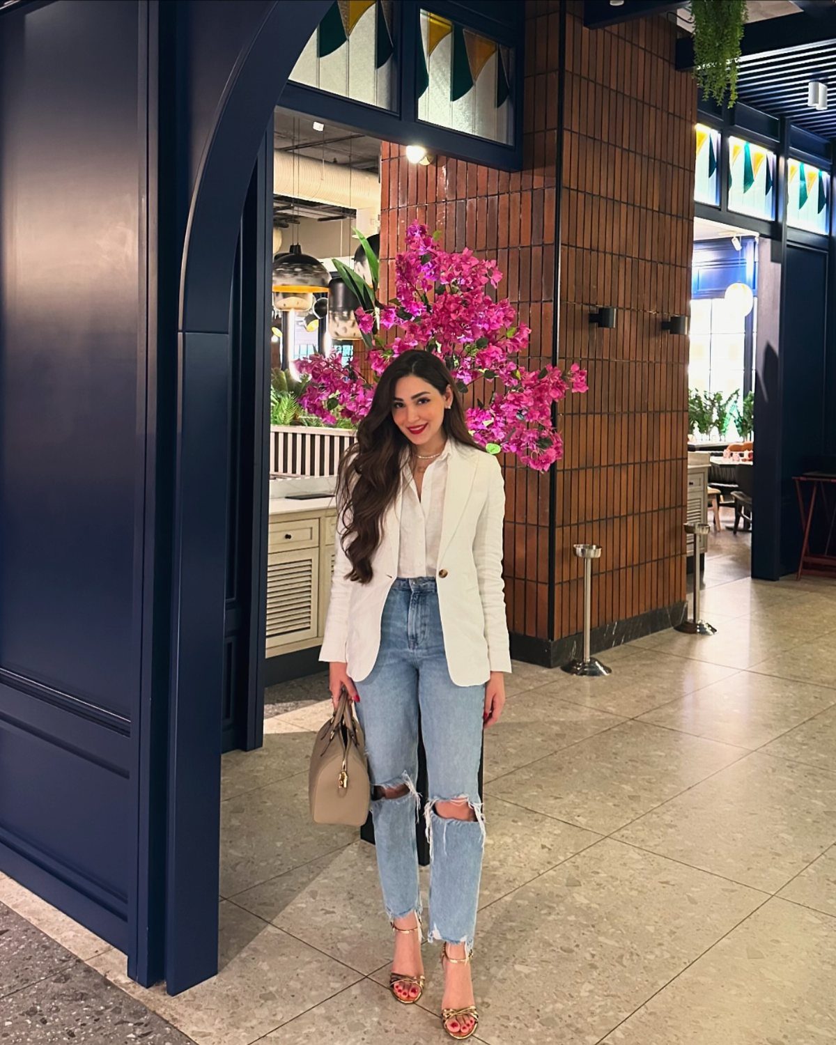 Woman posing in a white blazer, white shirt, and light blue ripped jeans with heels.