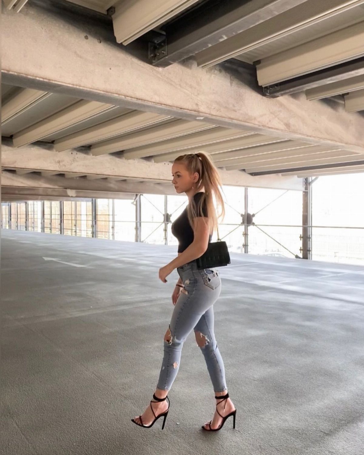 Woman walking in a parking garage wearing ripped jeans, a black crop top, and black high heels.