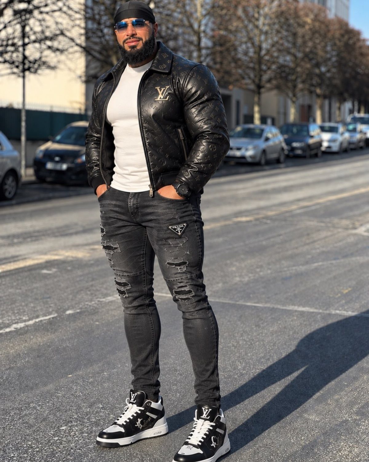 Man in a black leather jacket, white t-shirt, and grey ripped jeans standing on a street.