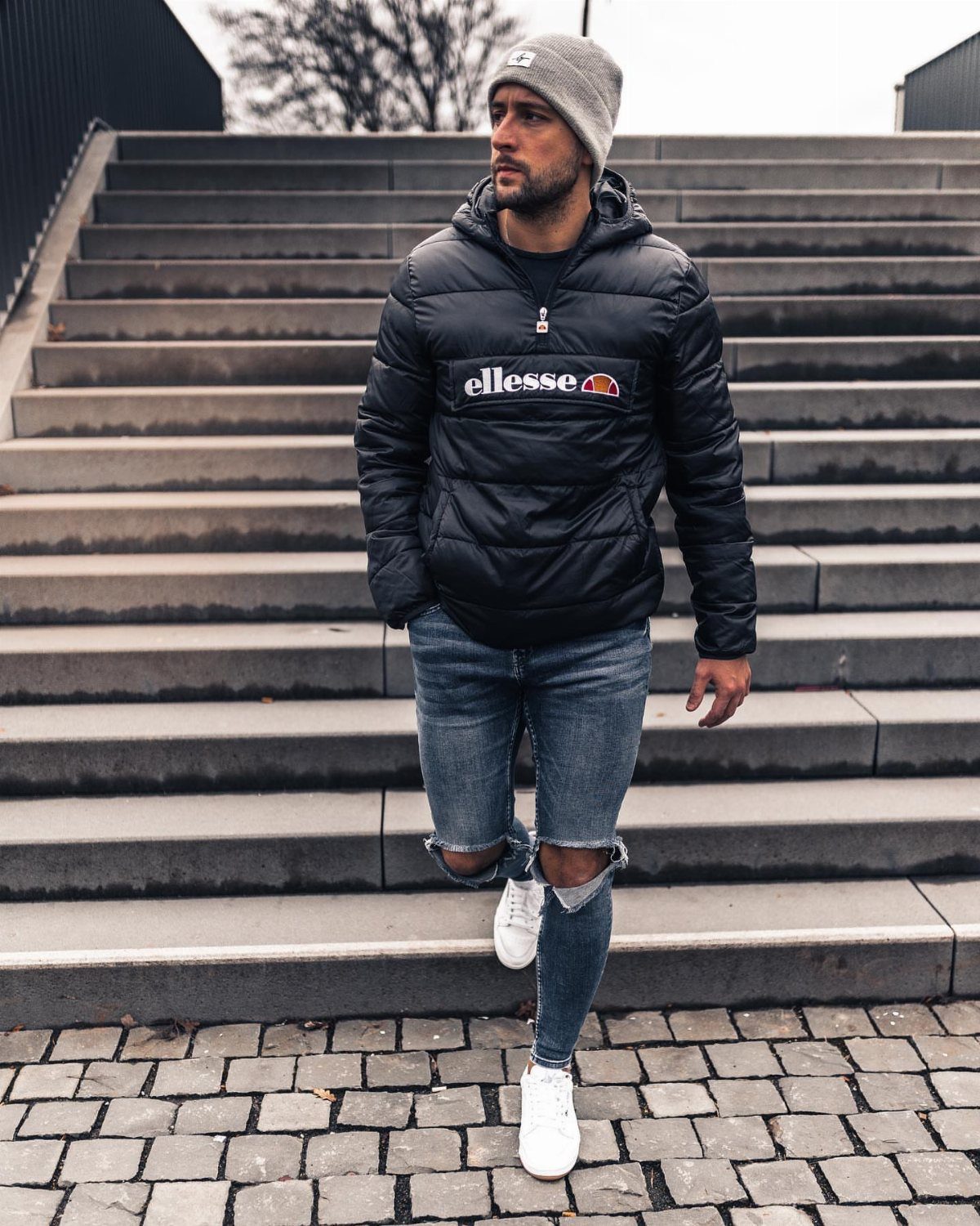 Man in a black puffer jacket and ripped jeans walking down outdoor stone stairs.