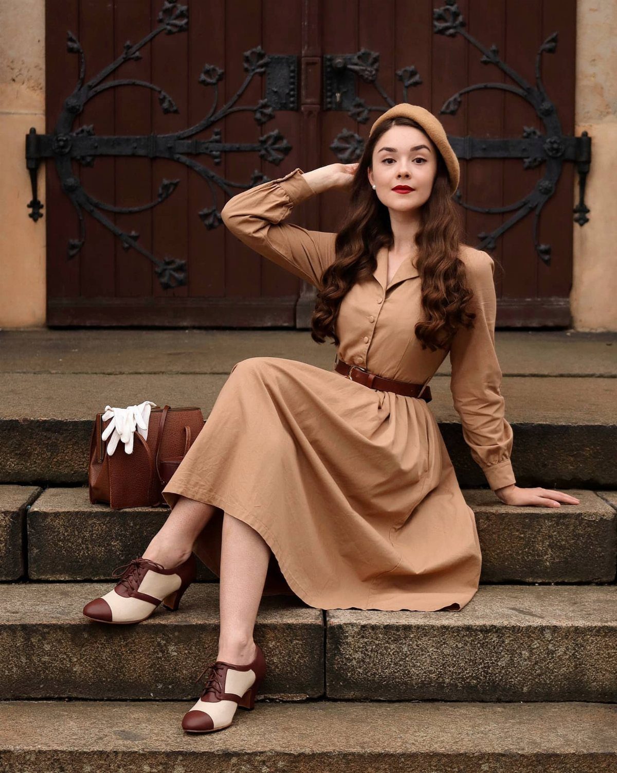 Woman in a tan midi dress and two-tone oxford shoes sitting on stone steps.
