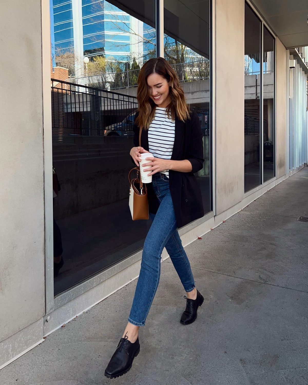 A woman walking outdoors wearing blue jeans, a striped shirt, and black oxford shoes.