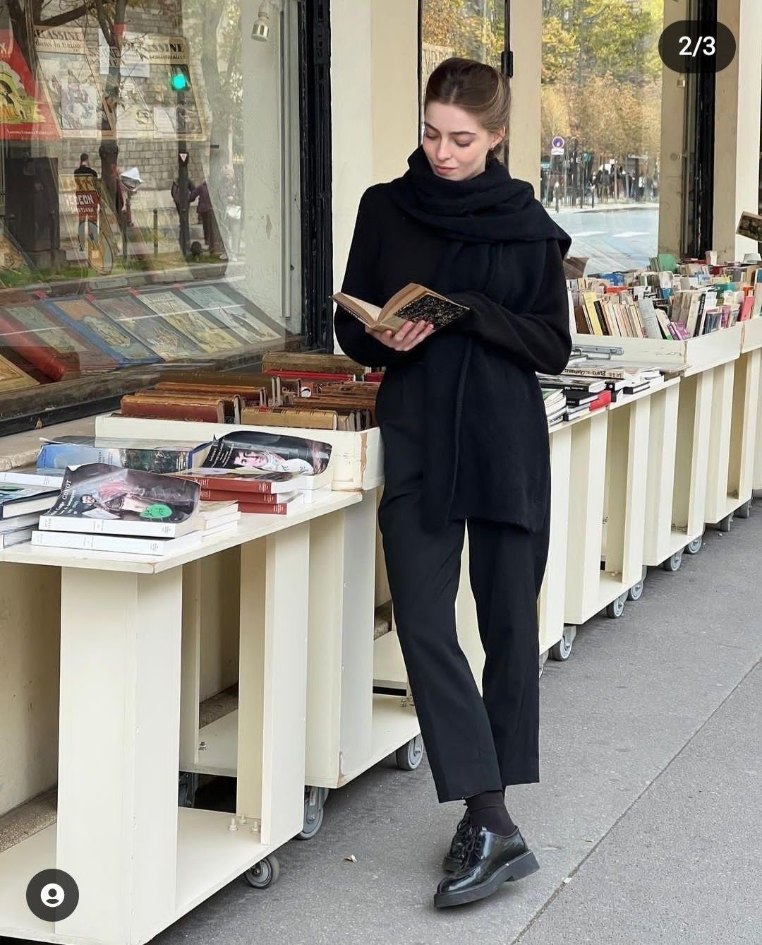 Woman in an all-black outfit including oxford shoes reading a book by a street bookstall.