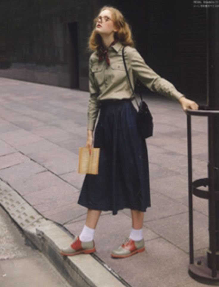 Woman wearing an olive shirt, long dark skirt, white socks, and tan oxford shoes standing outdoors.