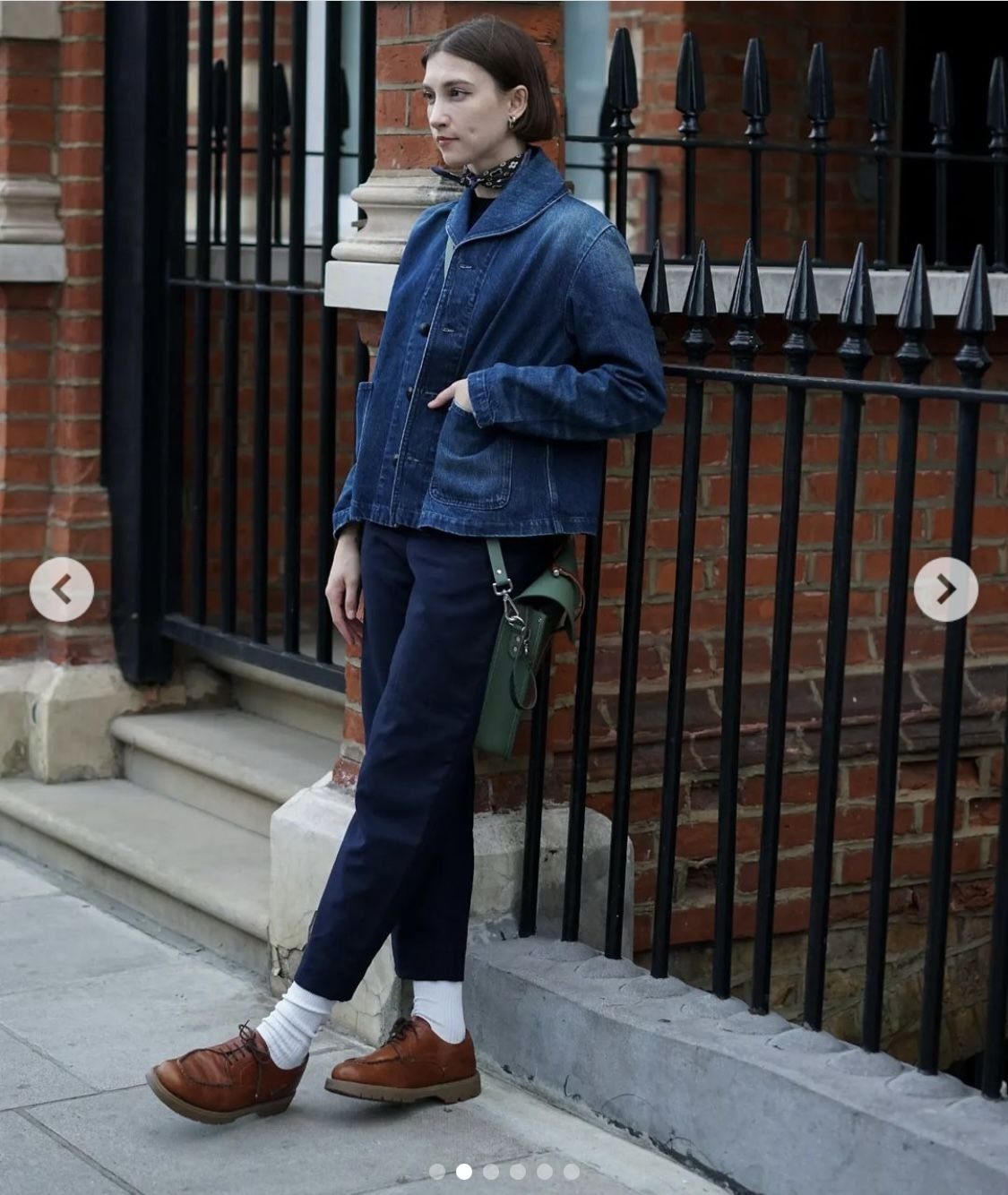 Woman wearing a blue denim jacket, dark trousers, white socks, and brown leather oxford shoes standing by a fence.