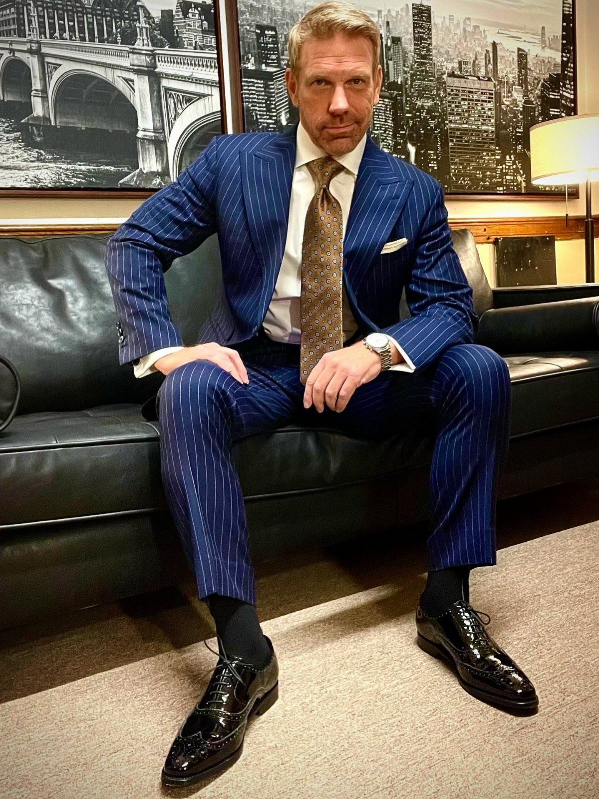 Man in a blue pinstripe suit sitting on a leather sofa wearing black wingtip oxfords.
