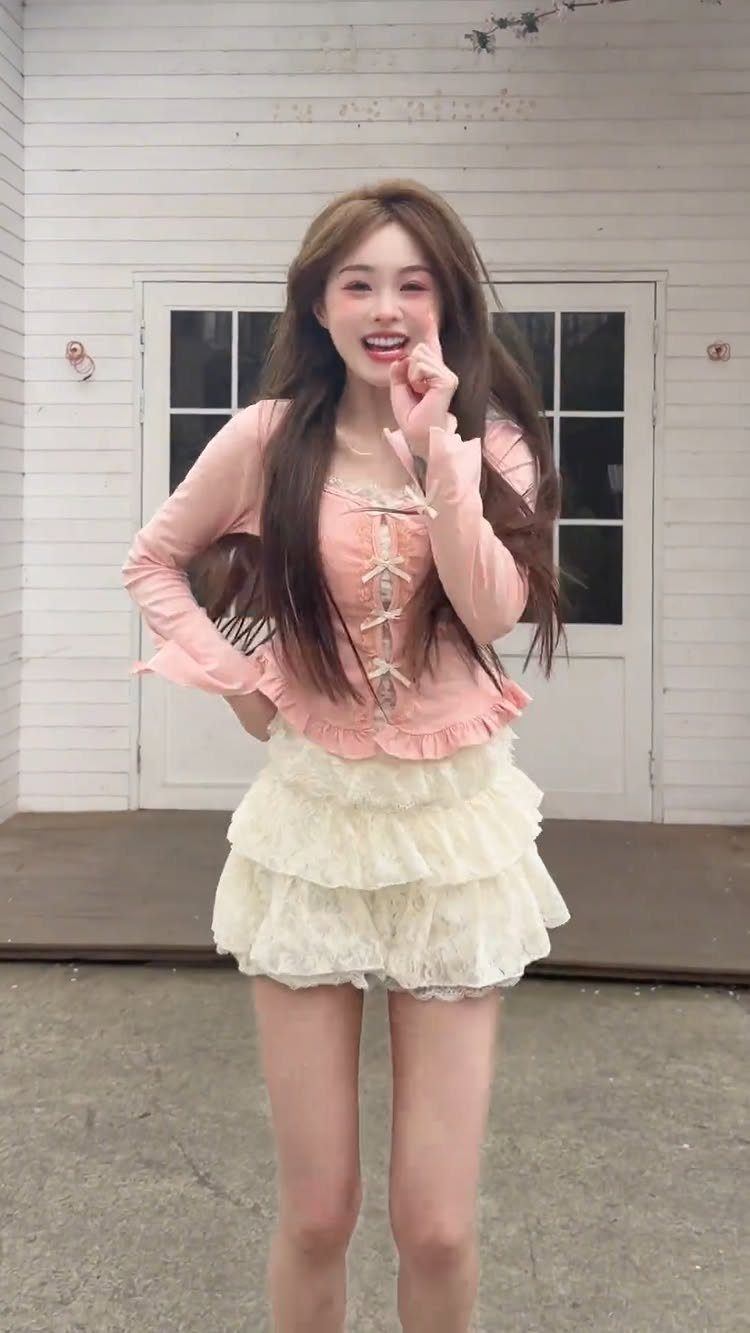 A smiling young woman wearing a pink corset top and a ruffled cream mini skirt.
