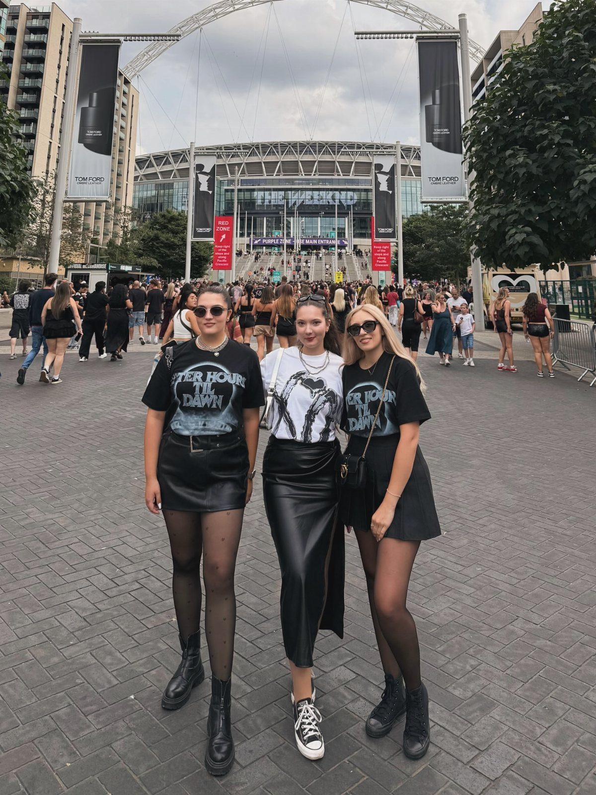 Three women posing in black graphic t-shirts, skirts, and boots in front of a large stadium.