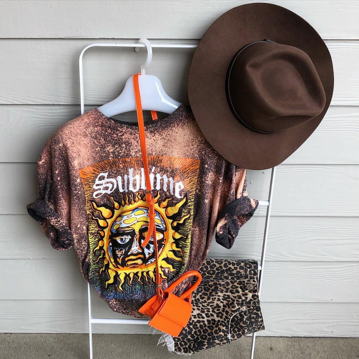 An oversized Sublime band t-shirt hanging on a rack with leopard print shorts and a brown hat.