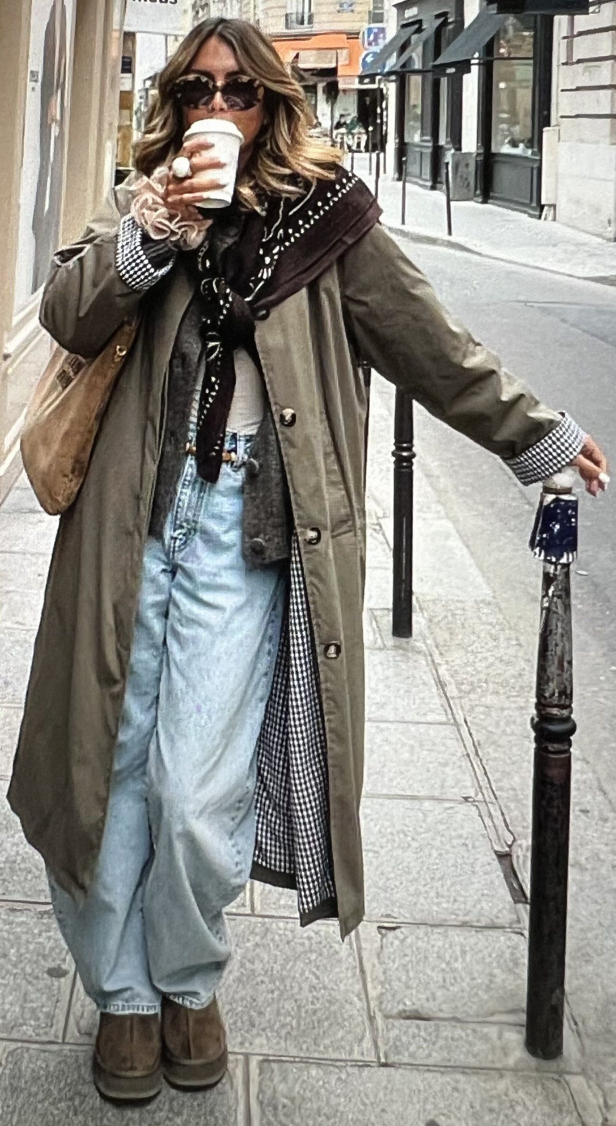 Woman in an olive trench coat, grey sweater, and light blue jeans standing on a city street.