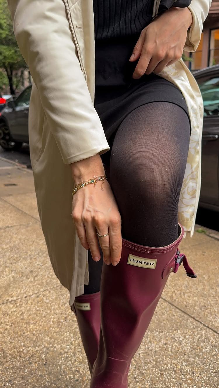 A person wearing a cream trench coat, black skirt, black tights, and maroon rain boots.