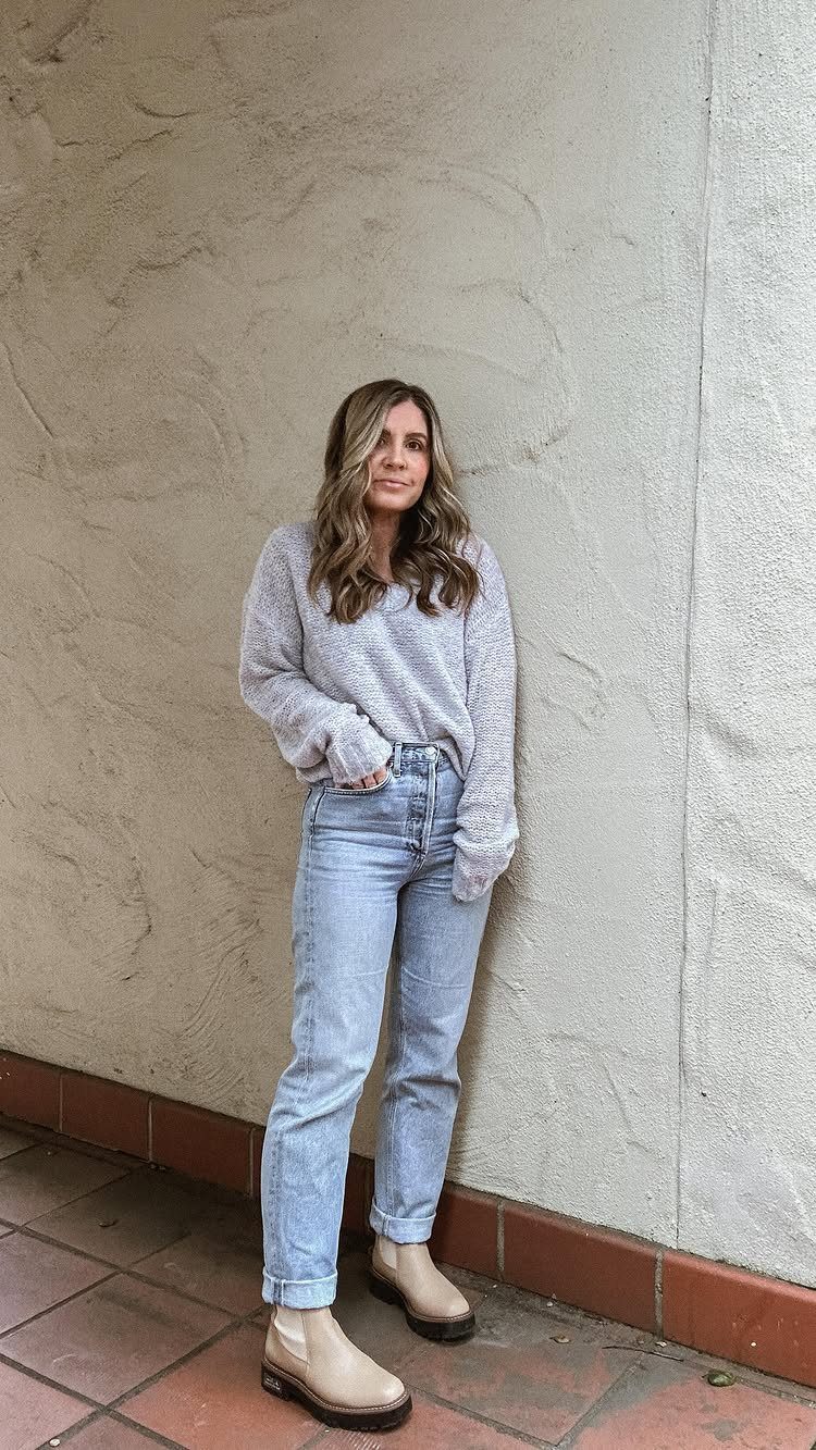 Woman wearing a grey sweater and light blue cropped jeans standing against a wall.