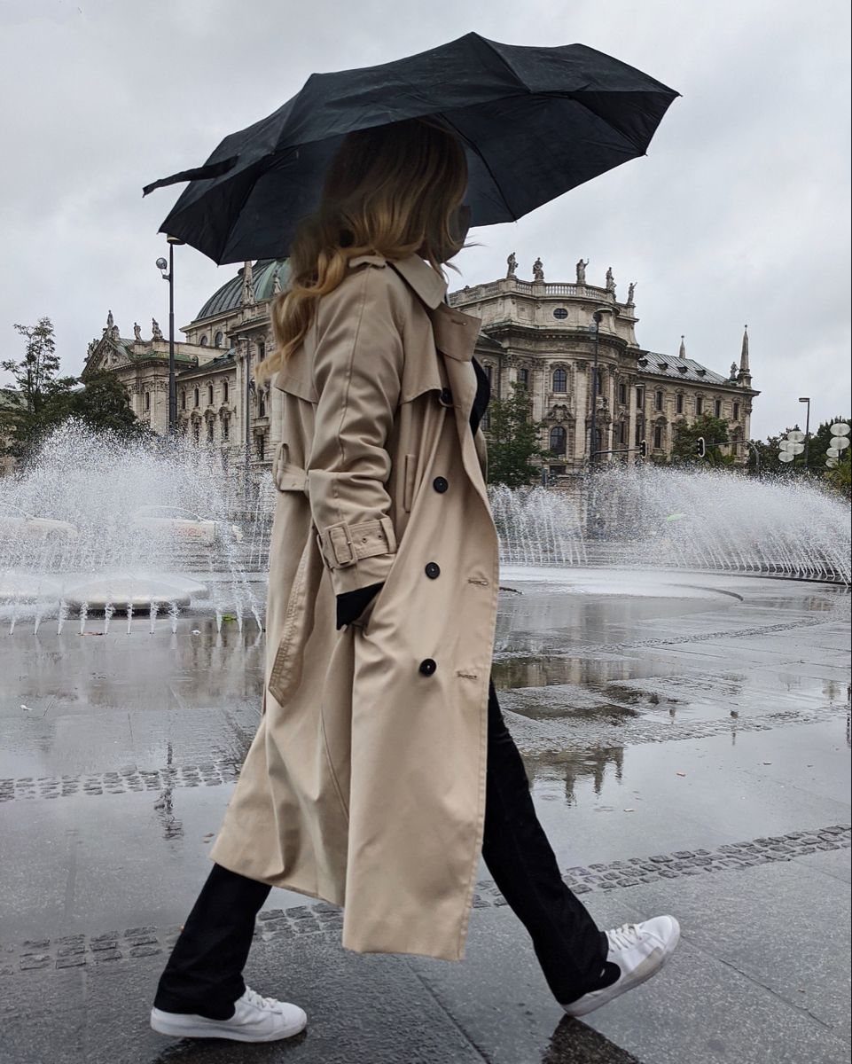 A woman in a beige trench coat and black slim pants walks past a fountain.