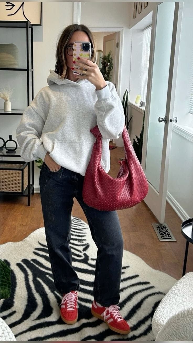 Woman wearing a grey hoodie, dark jeans, and red sneakers with a red handbag.