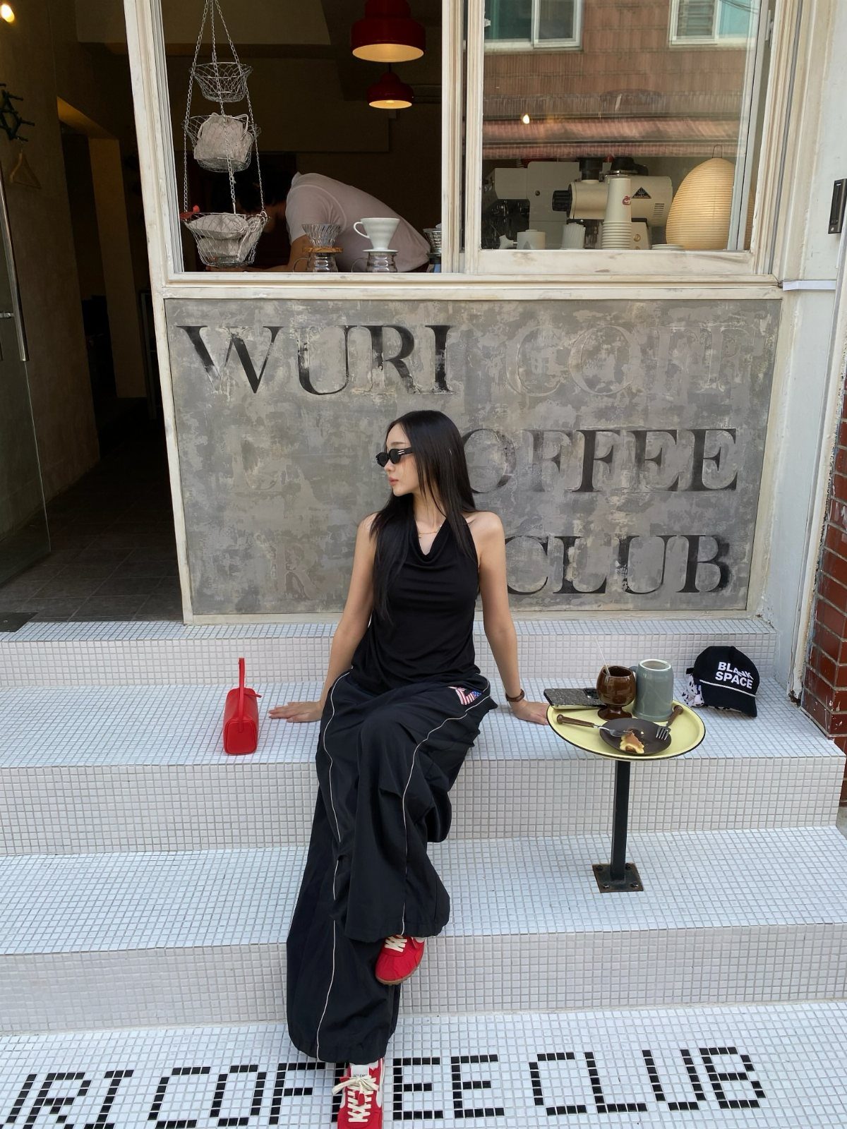 Woman in black outfit and red sneakers sitting on white tiled steps outside a coffee club.