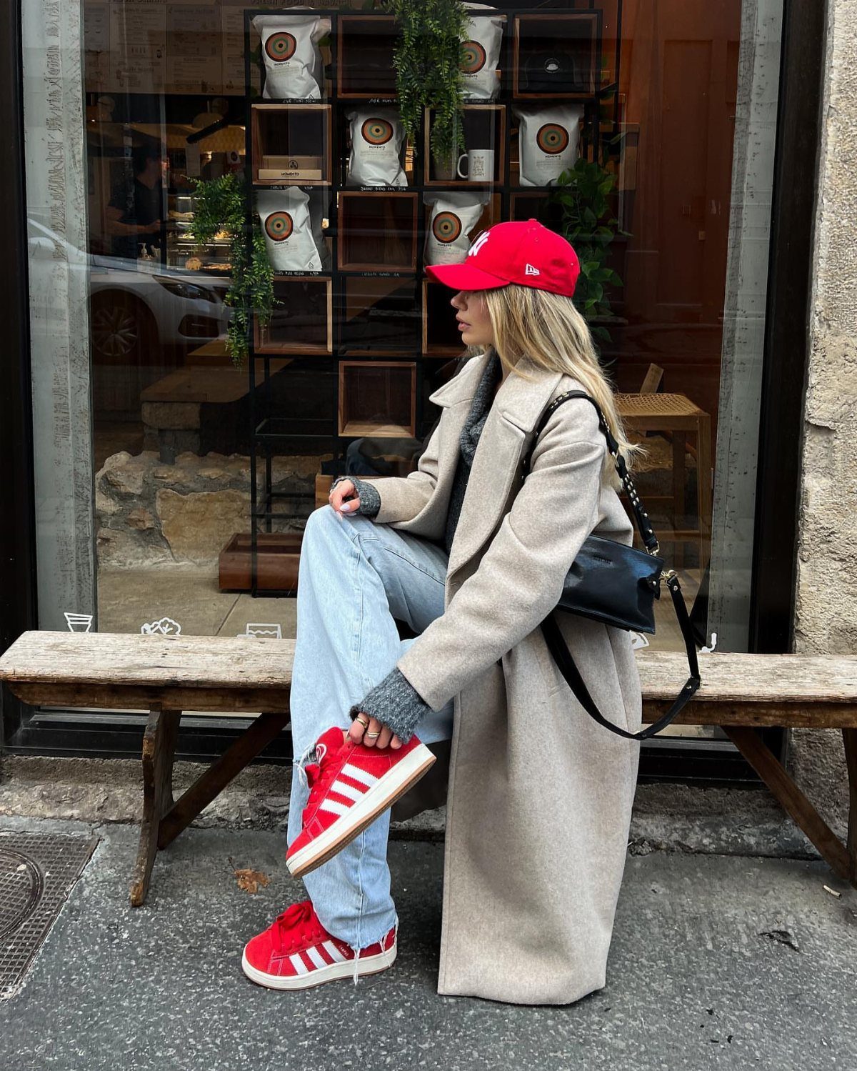 Woman in a beige coat and blue jeans wearing bright red sneakers and a red cap.