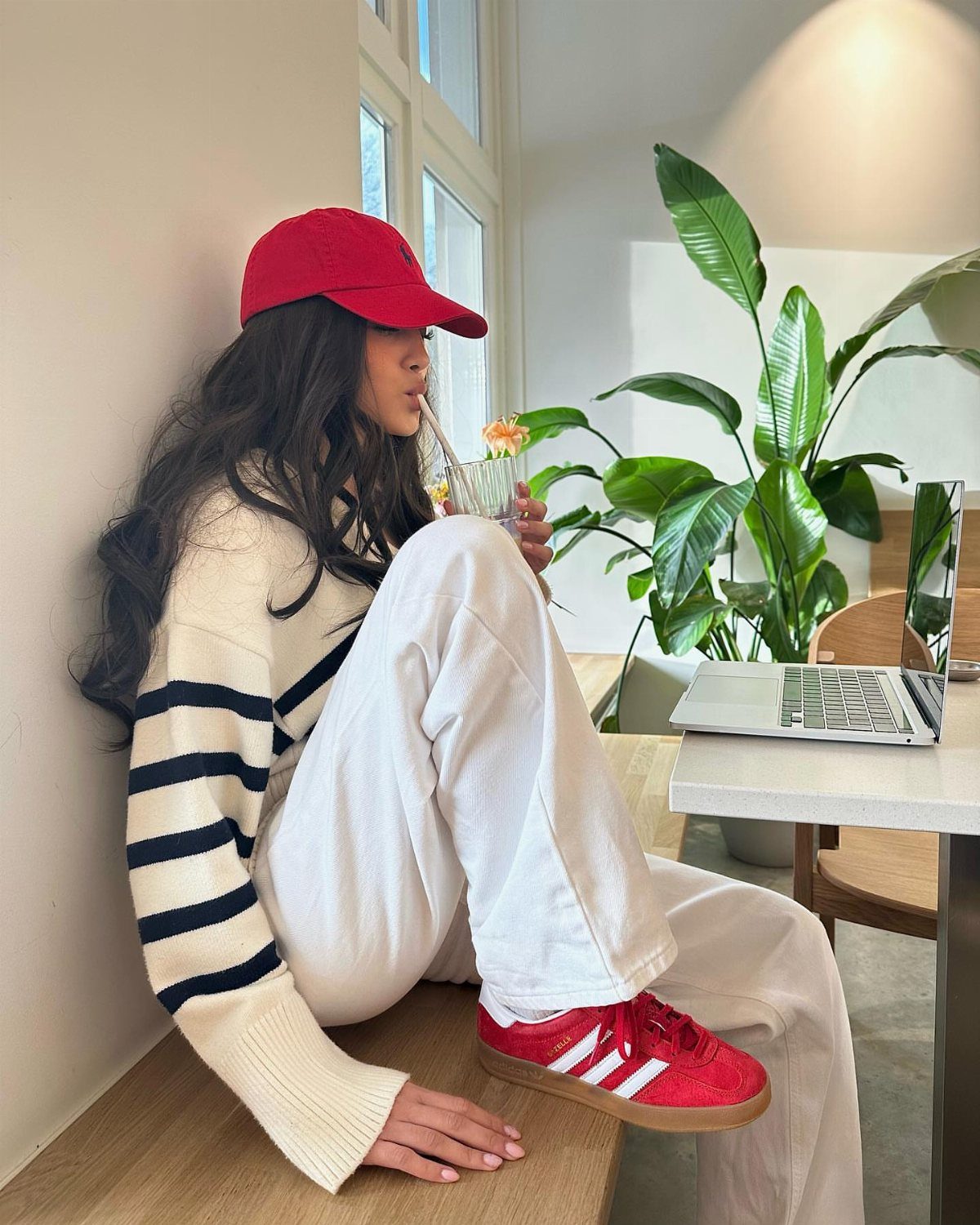 Woman in cream striped sweater and white pants wearing red sneakers and a red cap.