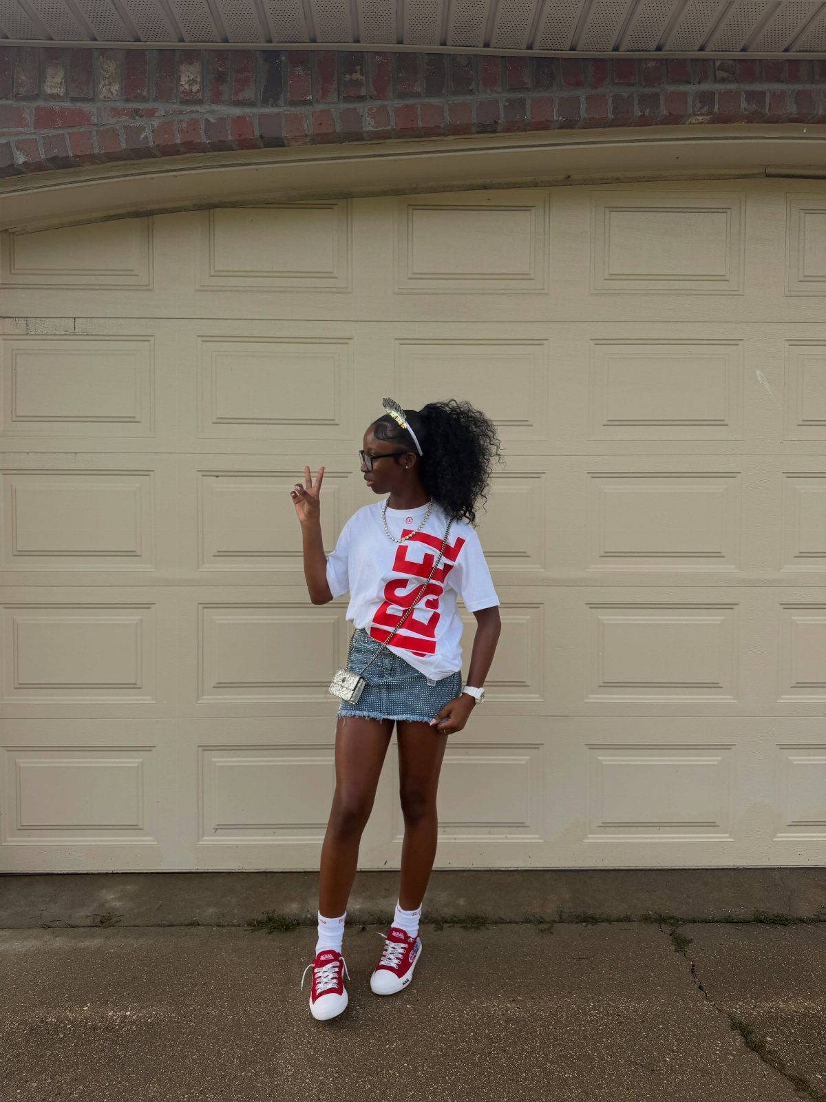 A young woman posing in a white t-shirt, denim shorts, and red sneakers.