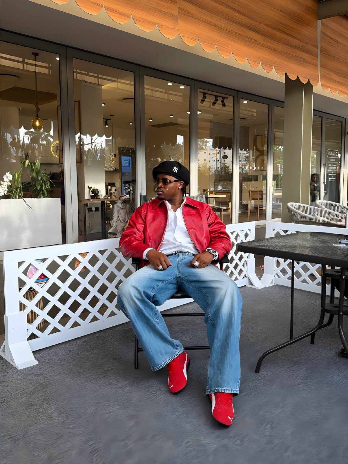 A man wearing a red leather jacket, blue jeans, and red sneakers sitting outdoors.