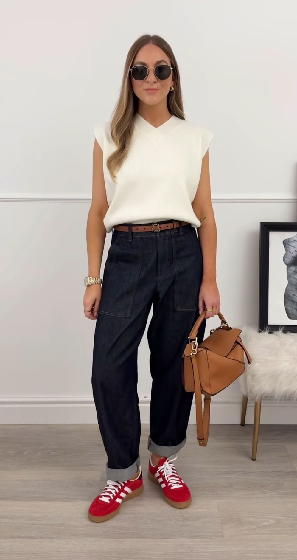 Woman posing in a white knit top, dark denim pants, and red sneakers.