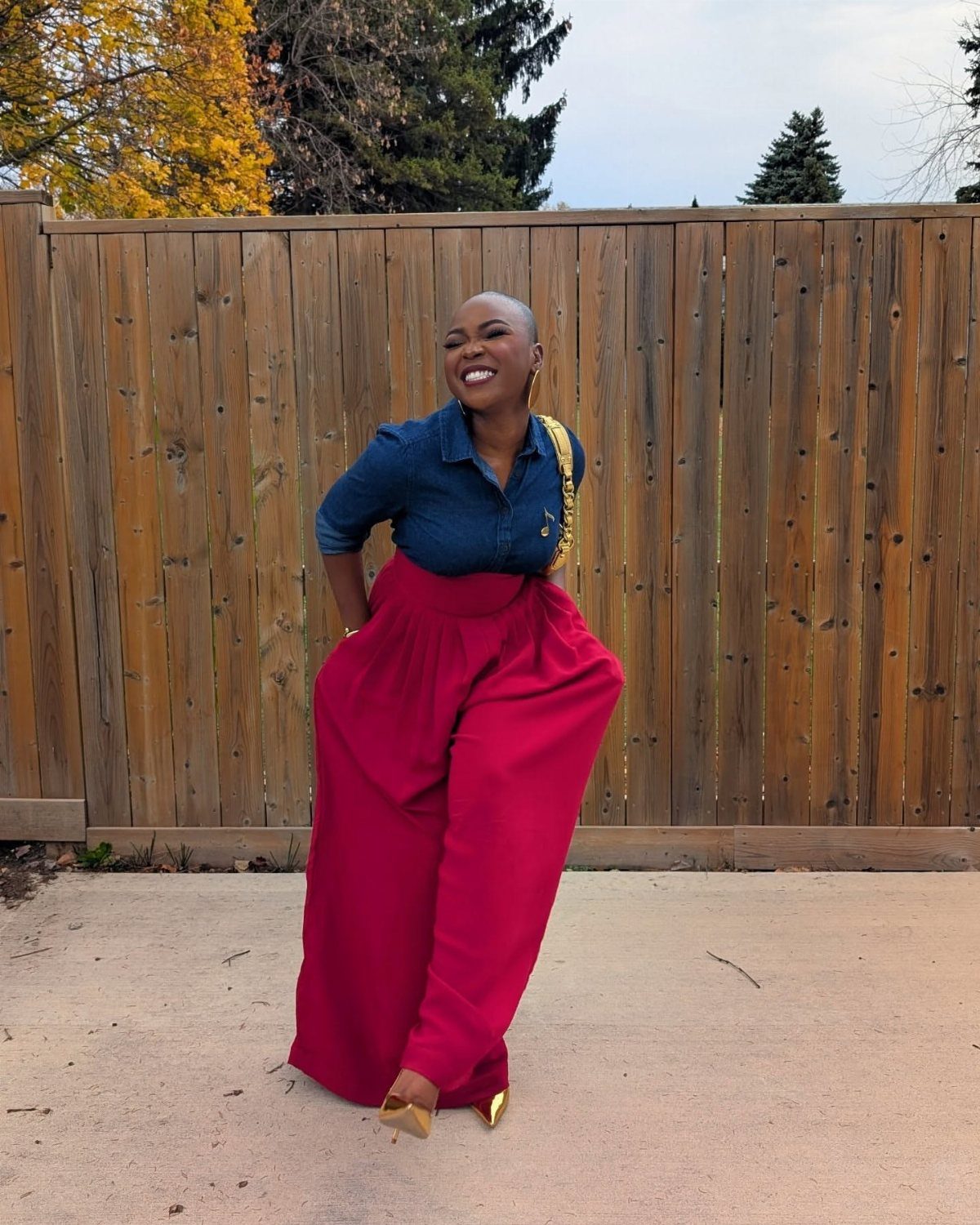 A woman smiling while wearing a blue denim shirt and bright red wide-leg trousers.