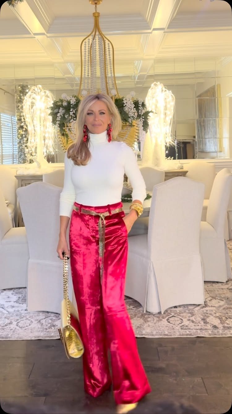 Woman wearing white turtleneck, red silk trousers, and gold accessories in a dining room.