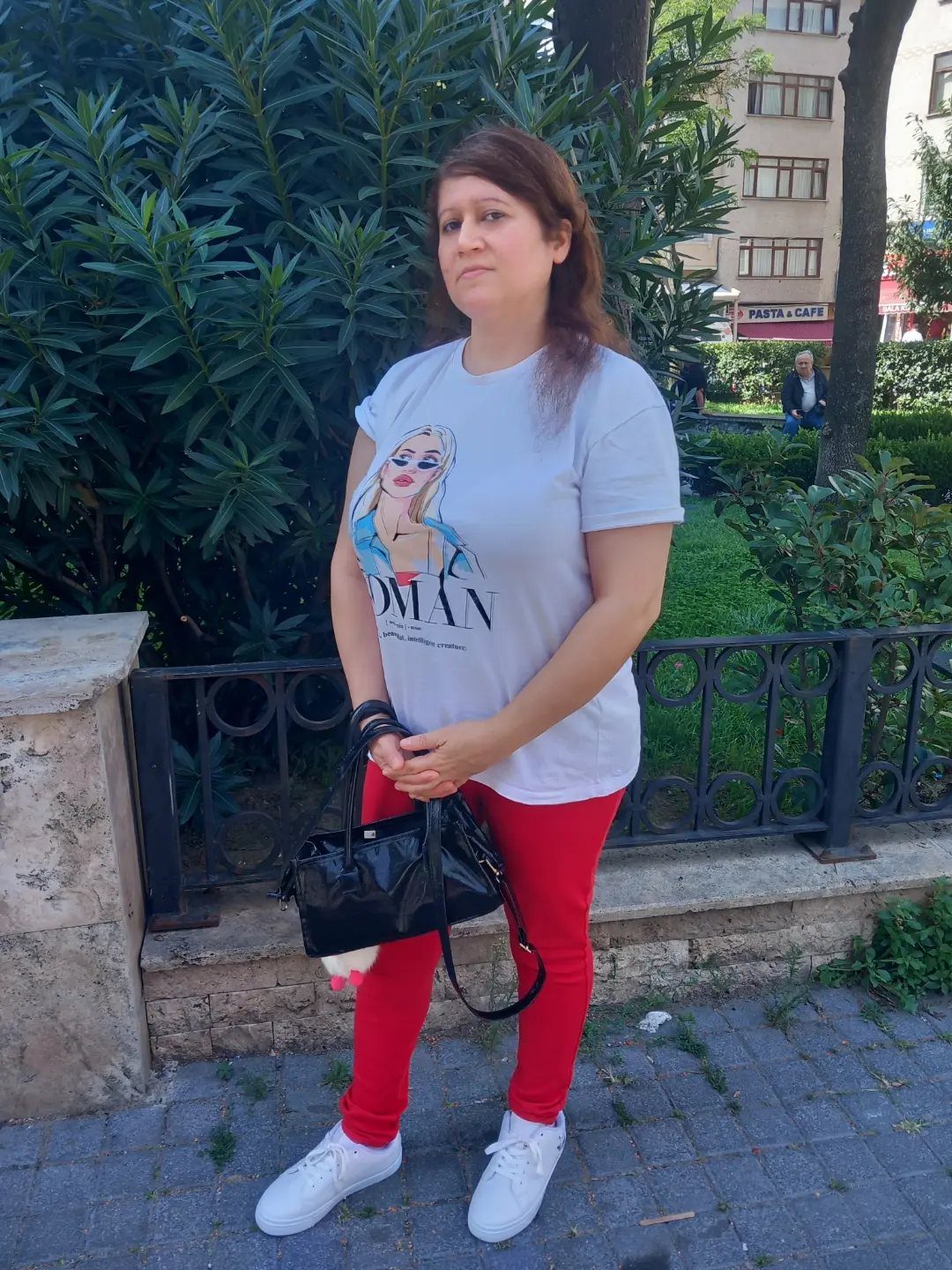 Woman wearing a white graphic t-shirt, red trousers, and white sneakers standing outdoors.