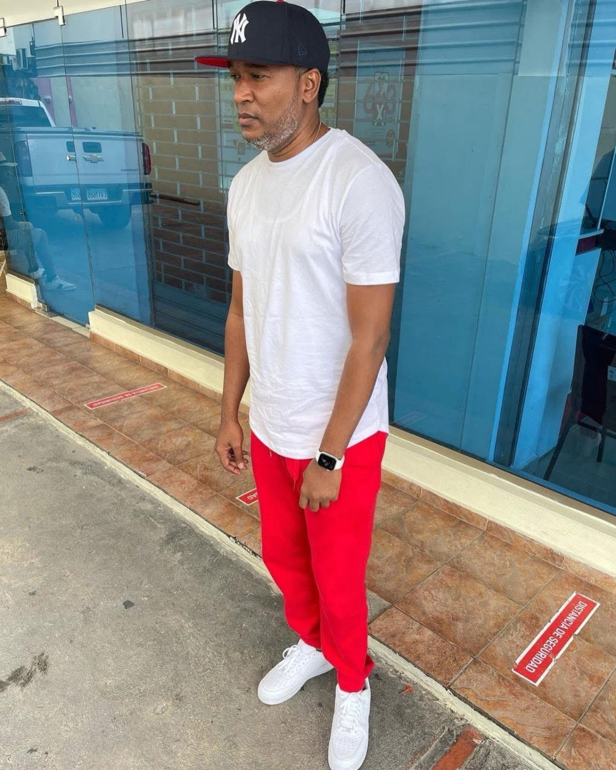 Man wearing a white t-shirt, red trousers, and white sneakers standing outdoors.