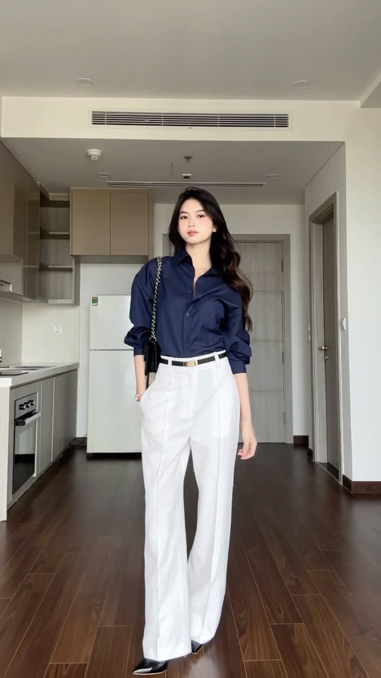 Woman wearing a navy blue button-down shirt and white wide-leg trousers standing in a room.