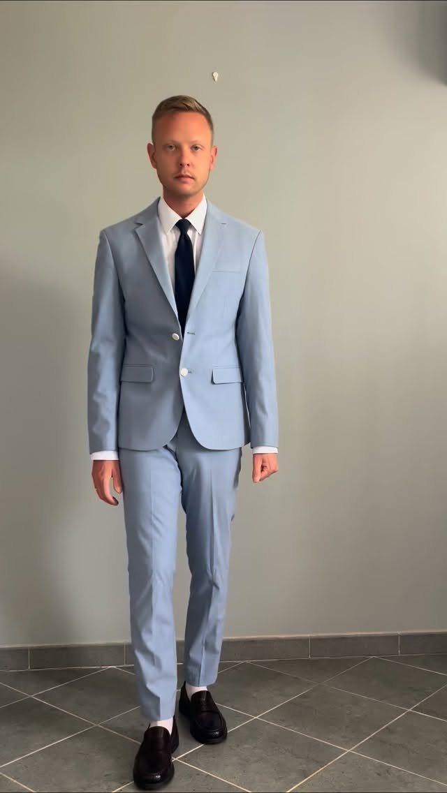 Man in a light blue suit and dark tie standing against a grey wall.