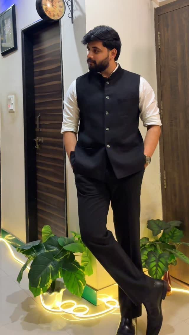 Man posing in a black waistcoat, white shirt, and black trousers indoors.