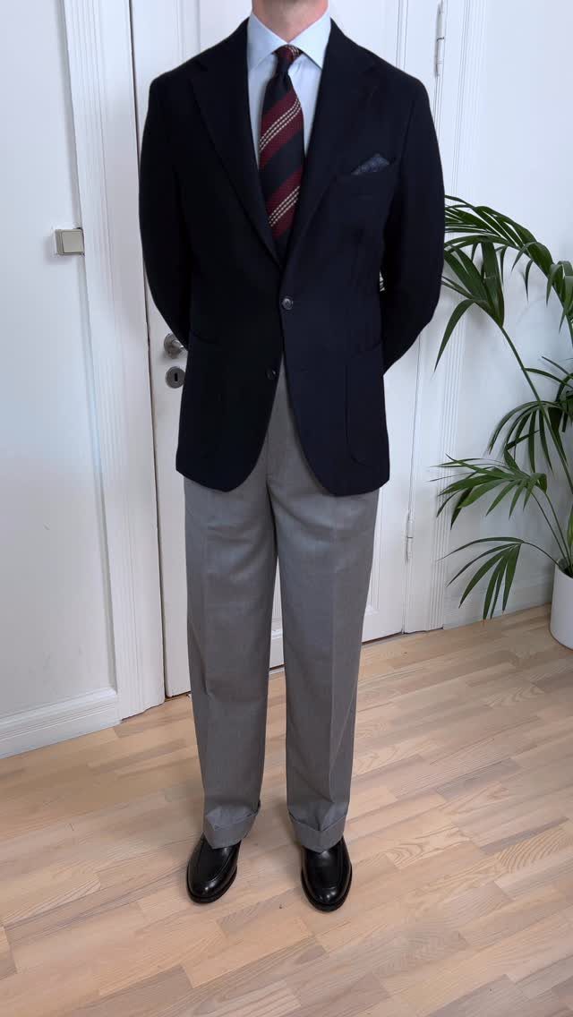 Man wearing a navy blazer, striped tie, light blue shirt, and grey trousers with black shoes.
