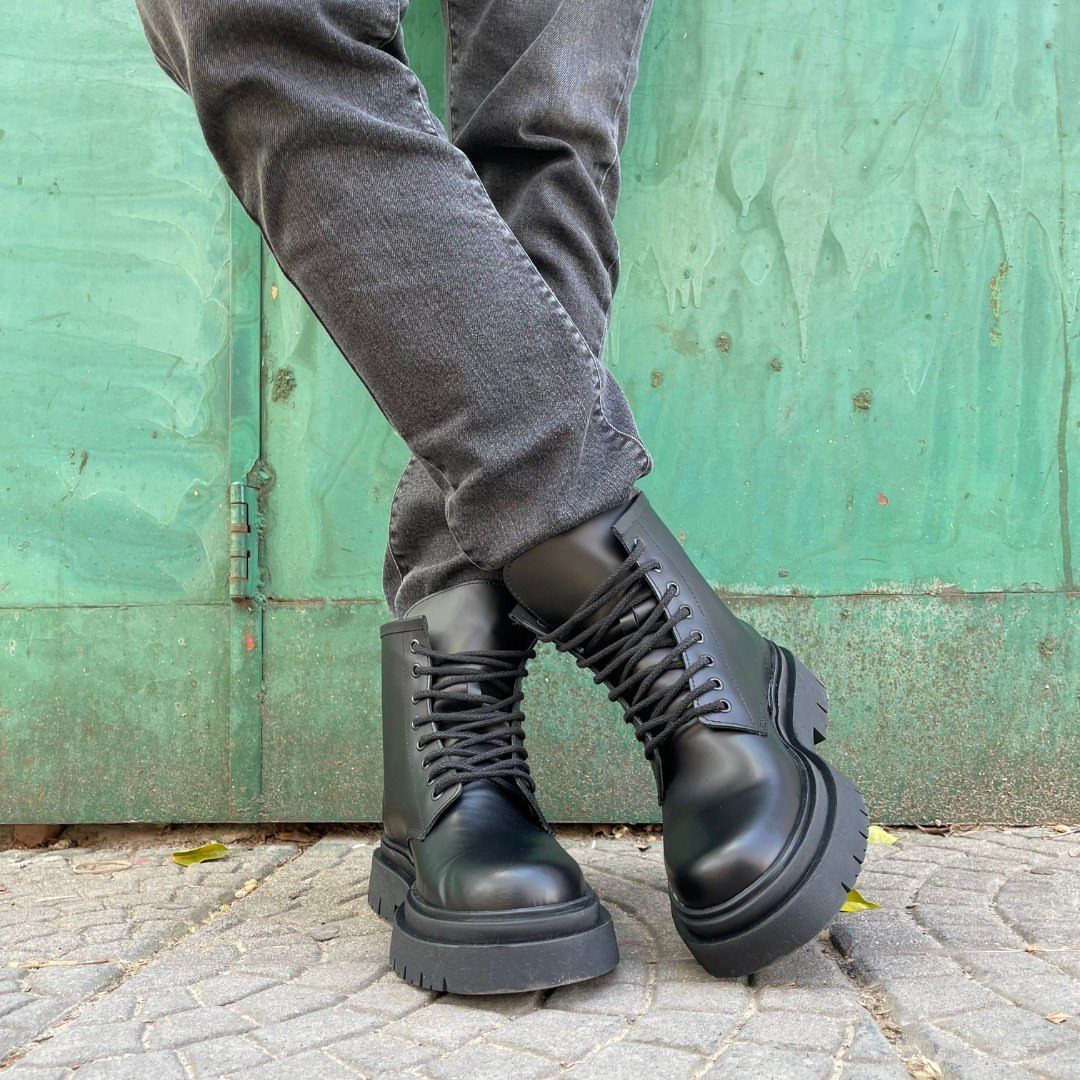 Black leather combat boots worn with dark grey jeans against a green wooden wall.