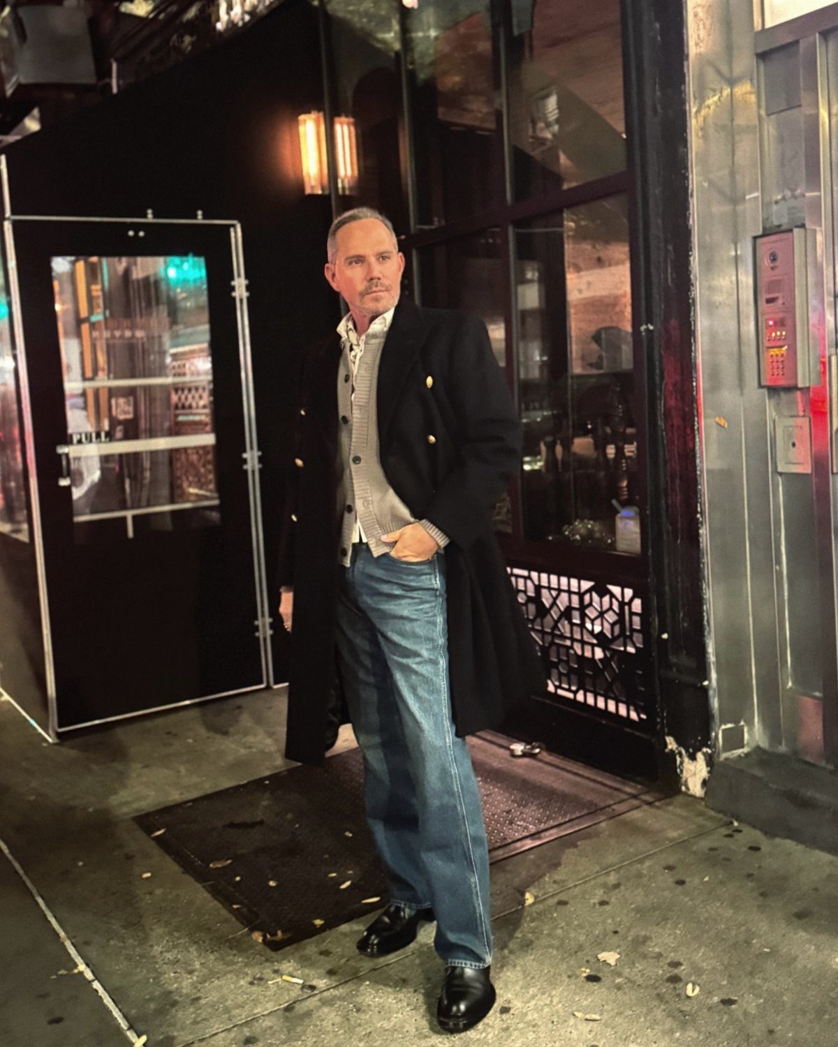 A man in a long black coat and blue jeans wearing polished black dress shoes.