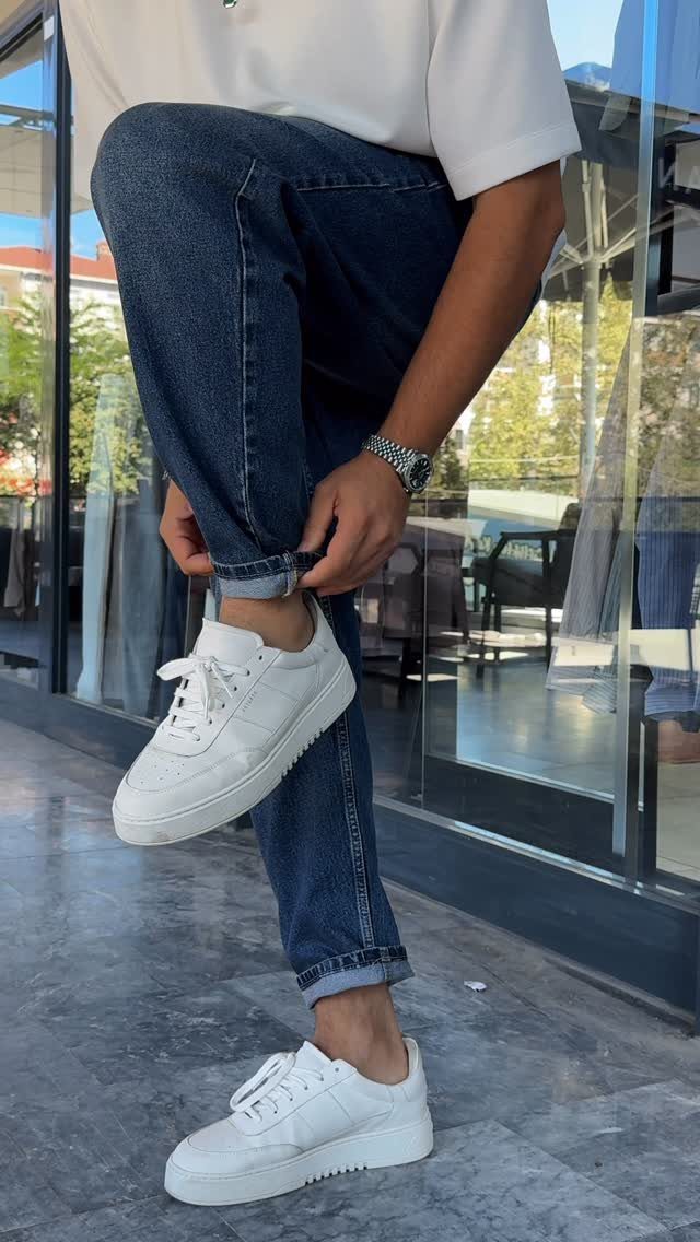 A person wearing dark blue jeans and white casual sneakers standing on a tiled floor.