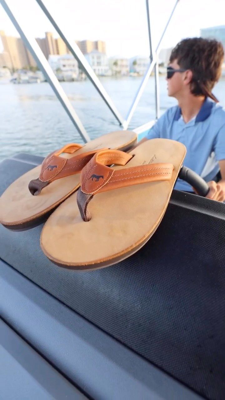 Tan leather flip-flops resting on the edge of a boat with a person in the background.