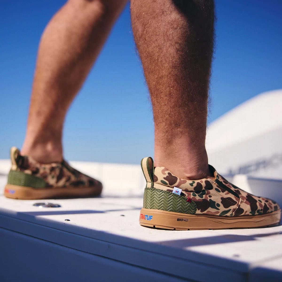 Person wearing camouflage slip-on shoes while standing on a white boat deck under a blue sky.