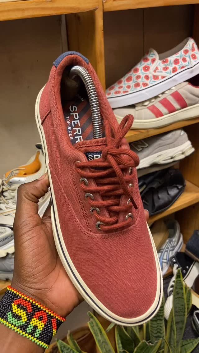 A hand holding a reddish-brown canvas sneaker in front of a wooden shoe shelf.