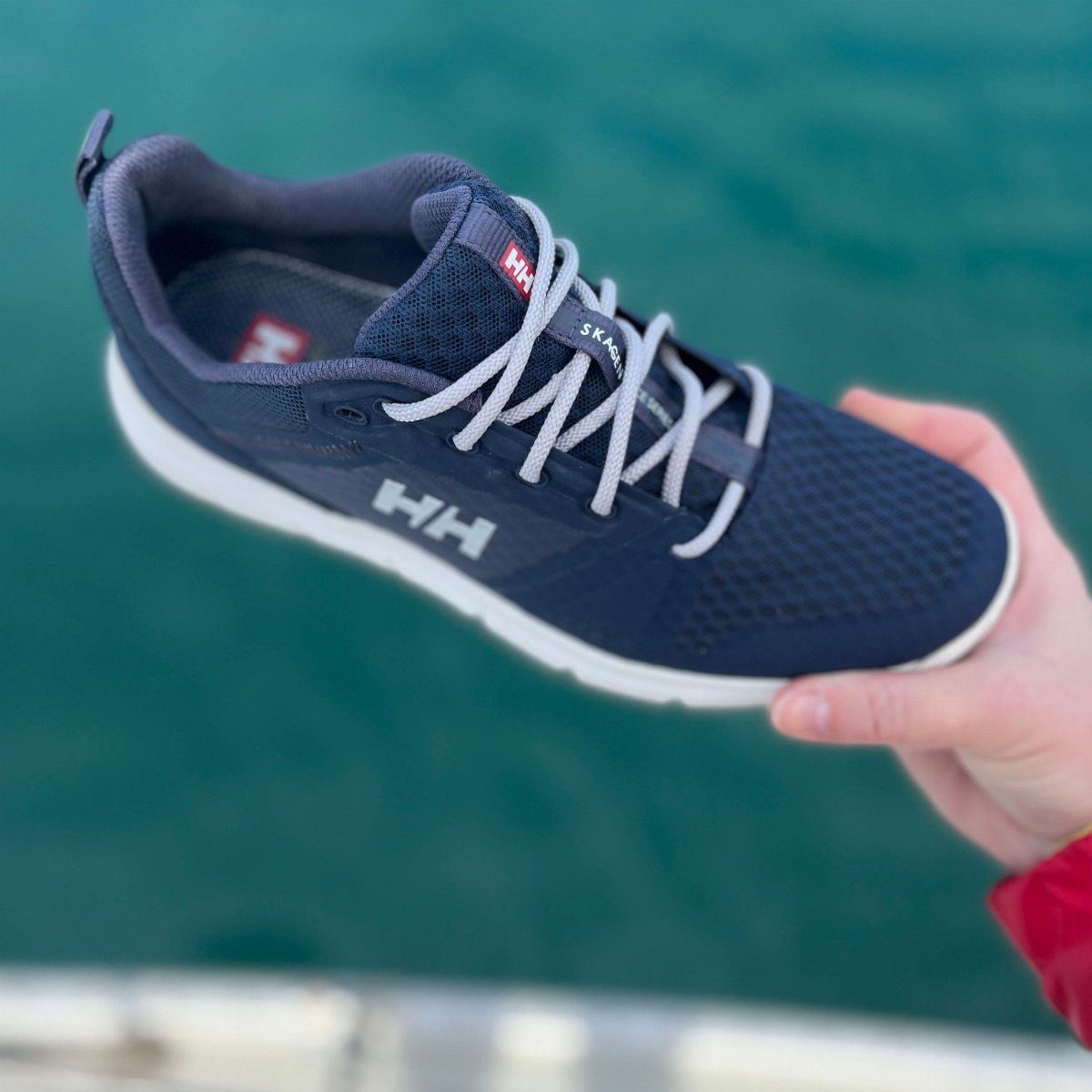 A hand holding a navy blue Helly Hansen Skagen sneaker over turquoise ocean water.