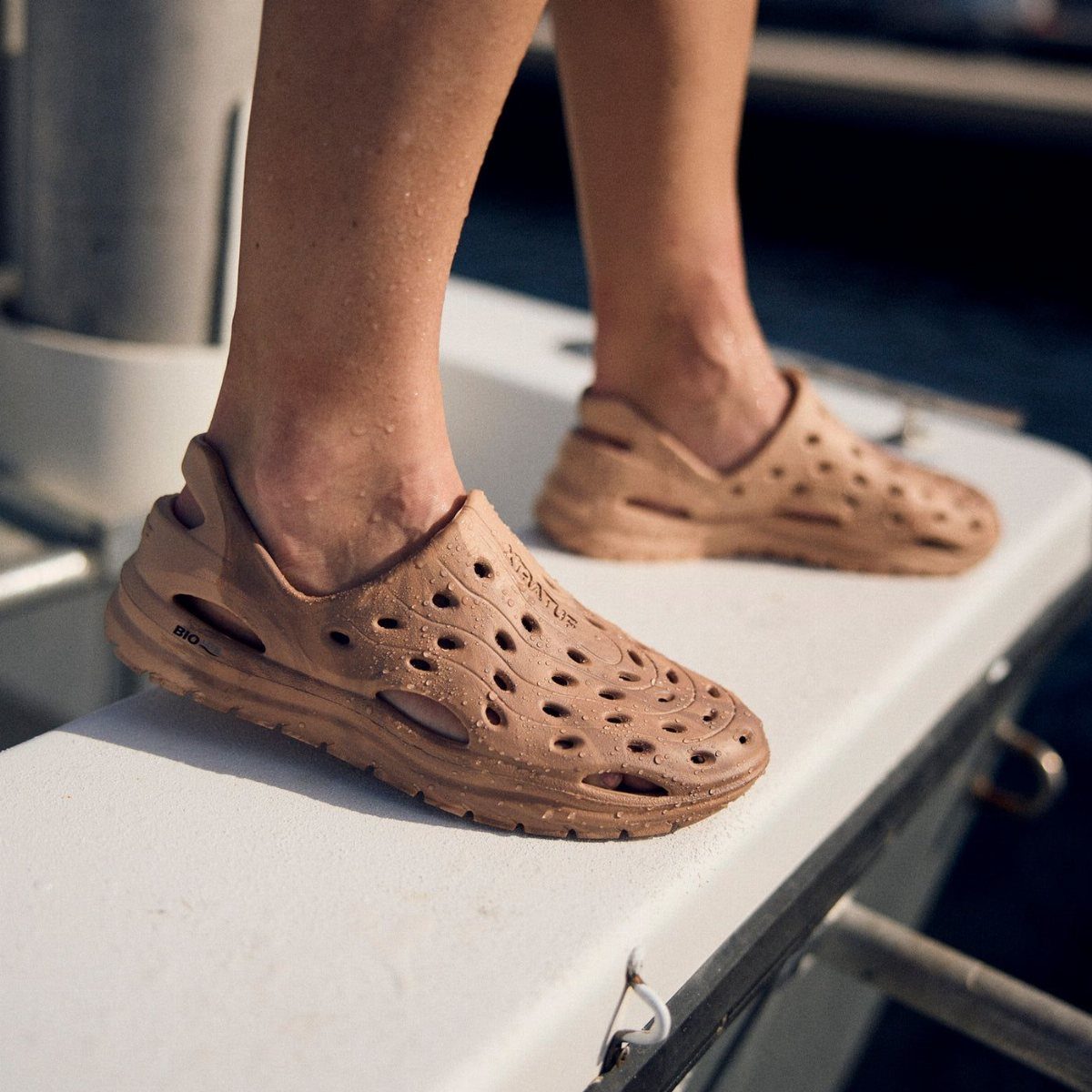 Tan perforated rubber slip-on shoes worn by a person standing on a white boat edge.