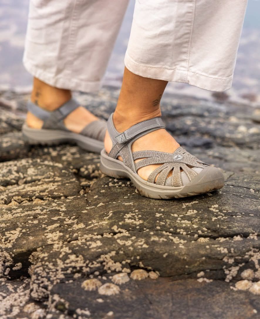 Grey sport sandals worn by a person standing on dark, rocky terrain near the ocean.